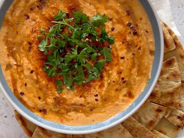 Creamy light orange dip in round bowl, garnished with cilantro, paprika and crushed red pepper. Pita chips on the side.