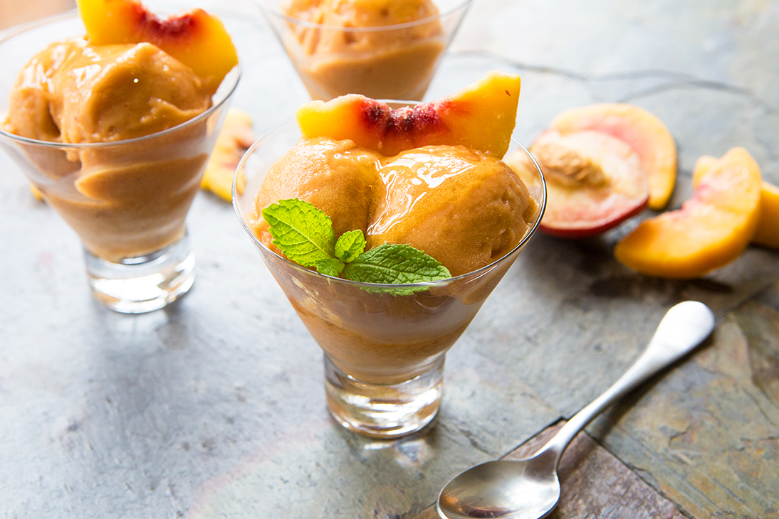 Peach sorbet served in glass cups, garnished with mint, on a textured stone background with peach halves.