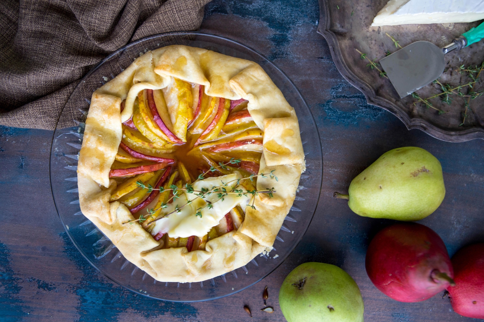 Rustic pear tart on a plate with pears and cloth on a blue wooden surface.