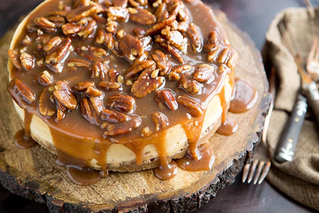 A caramel pecan honey cheesecake on a wooden stand with dripping caramel topping, next to cutlery on a burlap cloth.