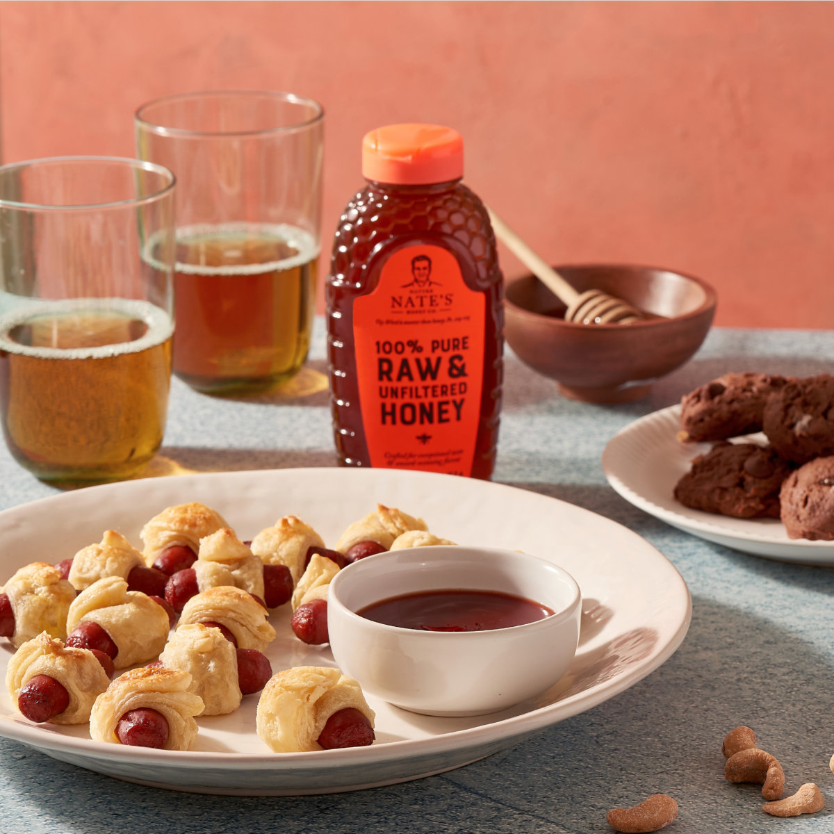Pigs in a blanket on a plate, cookies, a Nate’s honey bottle, with tea and wooden utensils, on a textured counter.