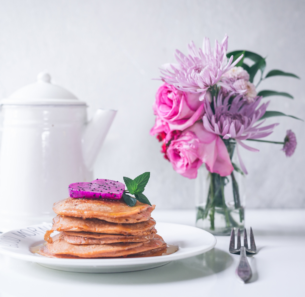 Stack of pancakes with a pink dragon fruit slice on top beside a bouquet and a teapot, elegant breakfast setup.