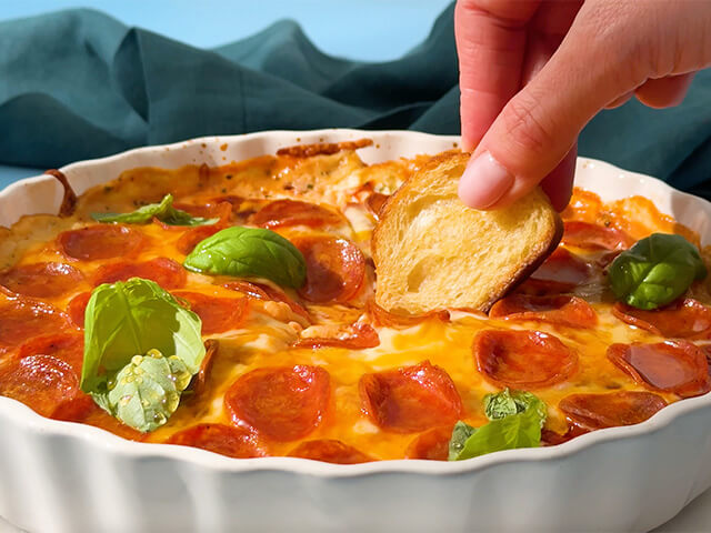 Hand dipping crostini into pizza dip that has pepperoni and basil as featured toppings.