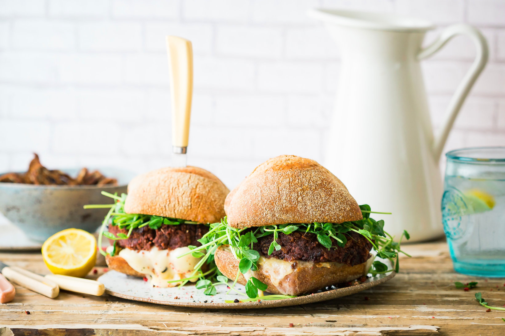 Two veggie burgers with pea shoots on a plate, with a rustic wooden background and a blue glass of water.