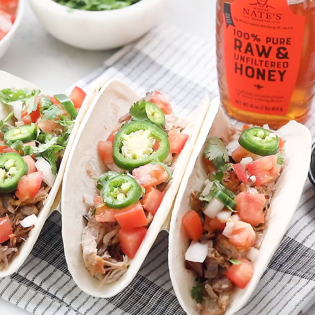 Slow cooker pork tacos with fresh toppings and a Nate's honey bottle in the background.