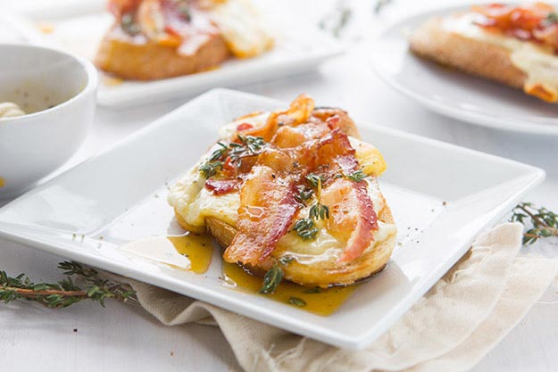 Open-faced sandwich with bacon and cheese, and drizzled with honey on a white plate, beige napkin beside, light setting.