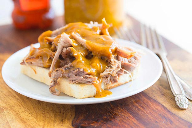 Pulled pork on toast with barbeque sauce on a white plate, silver fork on wooden table, sauce in background.