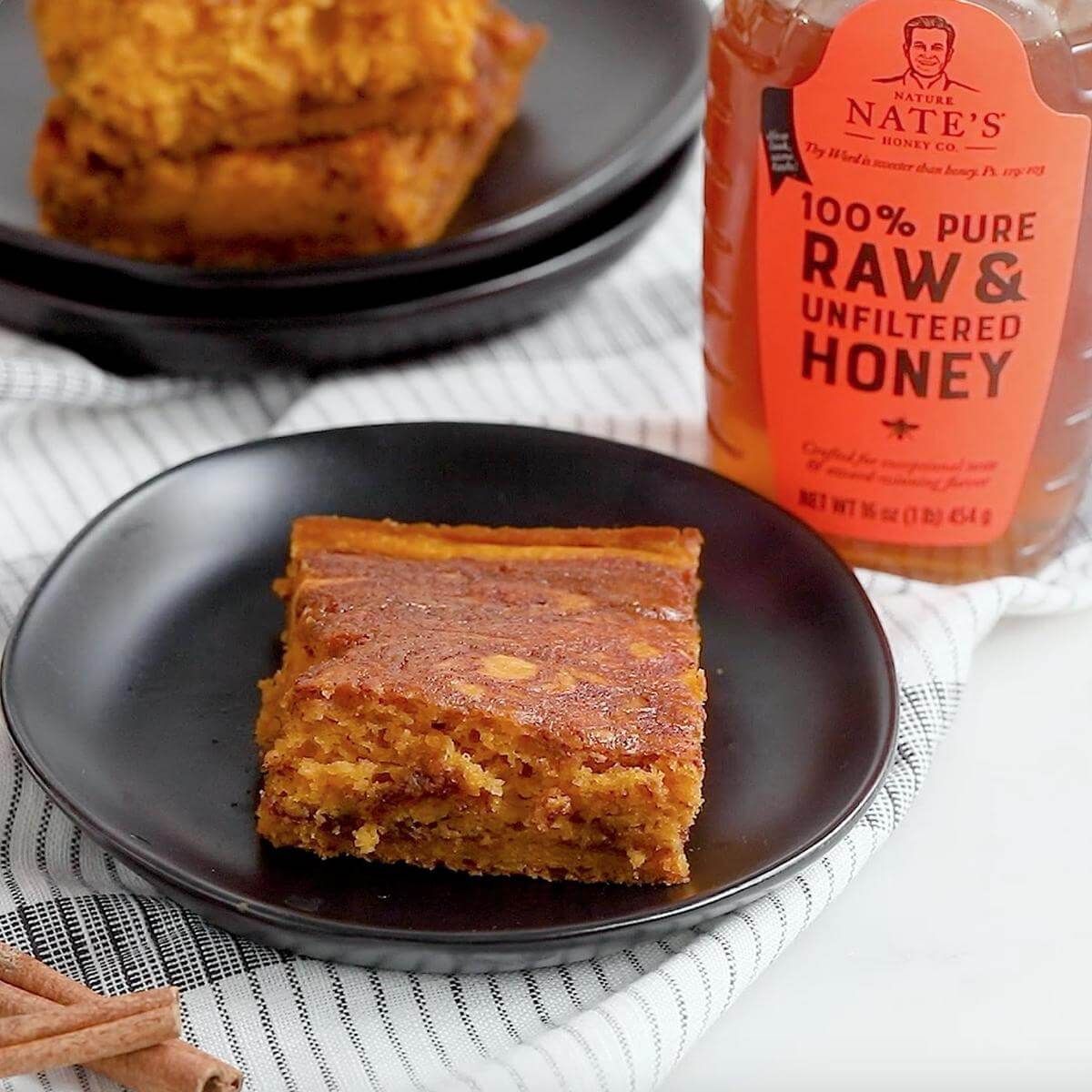 A slice of pumpkin coffee cake on a black plate with a bottle of Nate’s honey and cinnamon sticks on a checkered cloth.