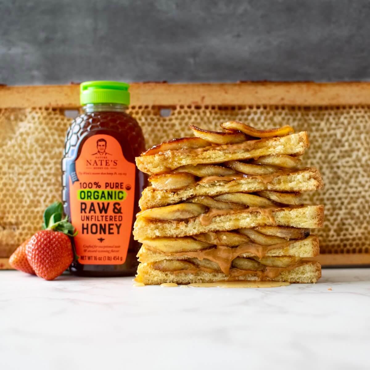 A stack of caramelized banana toast with a bottle of Nate’s organic raw & unfiltered honey and a strawberry.