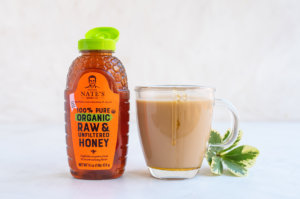 Nature Nate's organic honey bottle next to a cup of tea and fresh leaves on a white background.