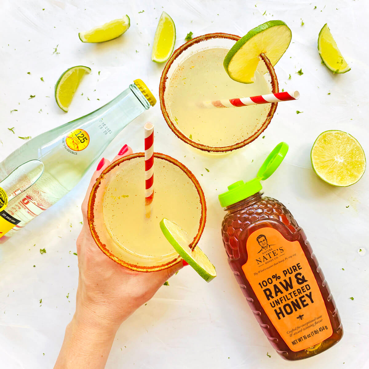 Hand holding a margarita, bottle of soda, bottle of Nate’s organic honey and  lime wedges on white surface.