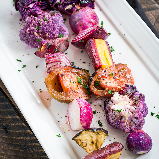 Two skewers of grilled purple vegetables on a white platter, wooden table backdrop.