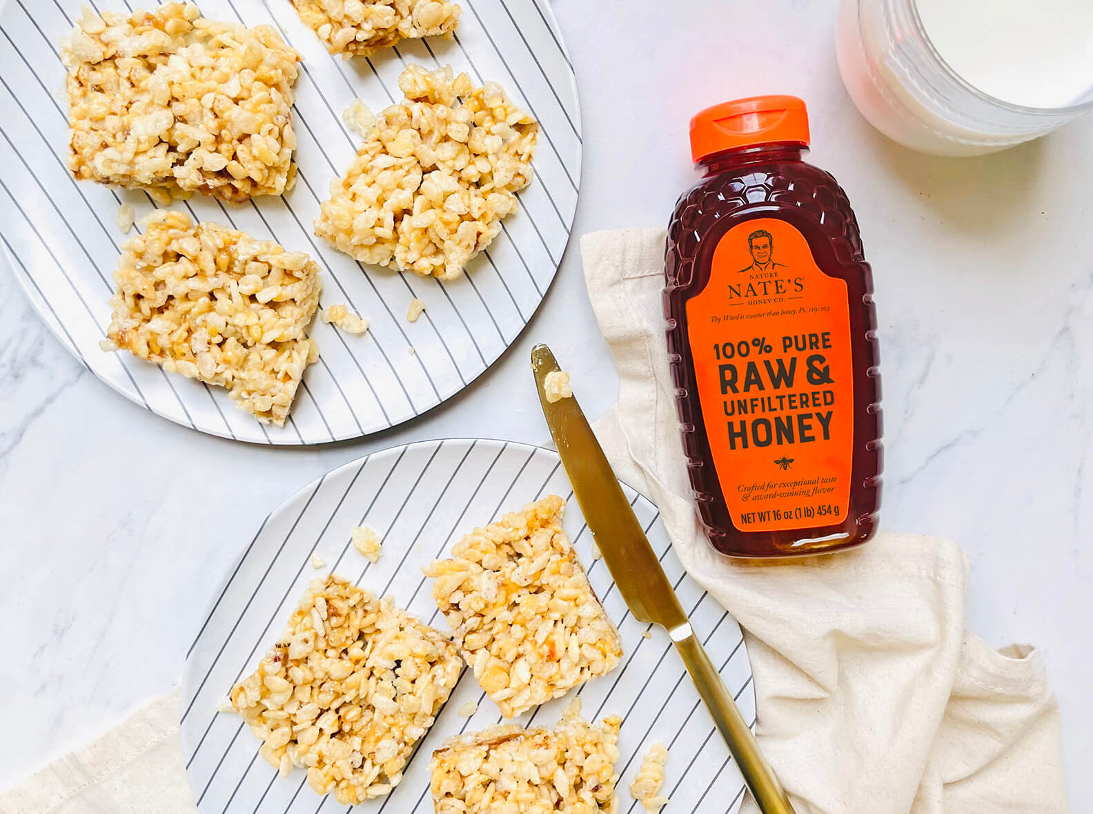 Rice crispy treats made with honey on a striped plate, a bottle of Nate’s honey, and a glass of milk on marble.