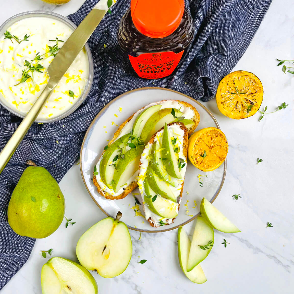 Toast with ricotta and pear slices, a bowl of whipped ricotta, and a bottle of Nate’s honey on a marble tabletop.