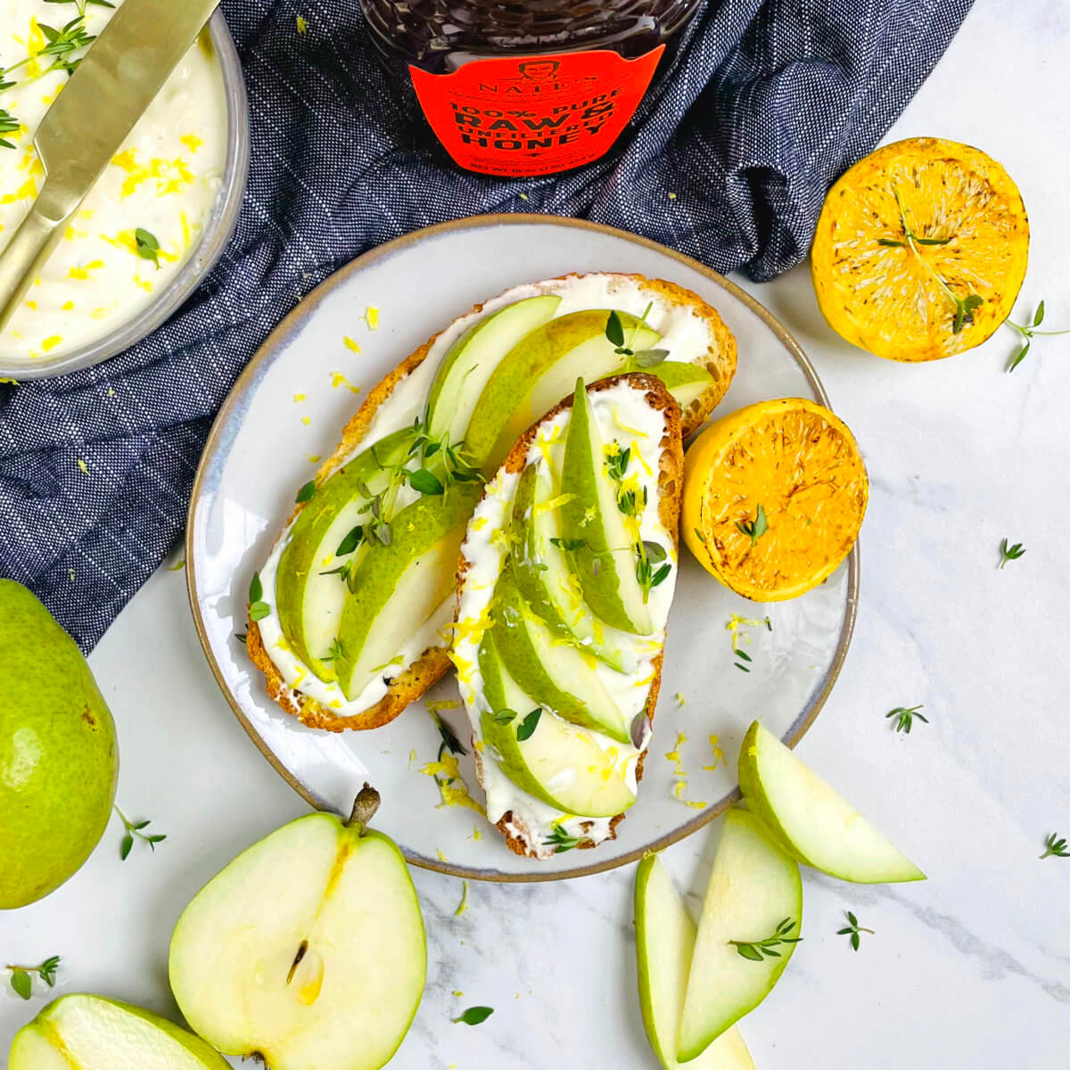 Toast with ricotta and pear slices, a bowl of whipped ricotta, and a bottle of Nate’s honey on a marble tabletop.