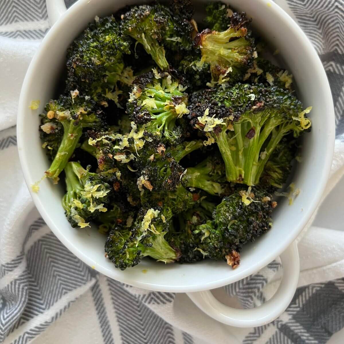 Roasted broccoli garnished with lemon zest in a white casserole dish with handles, on a grey and white cloth.