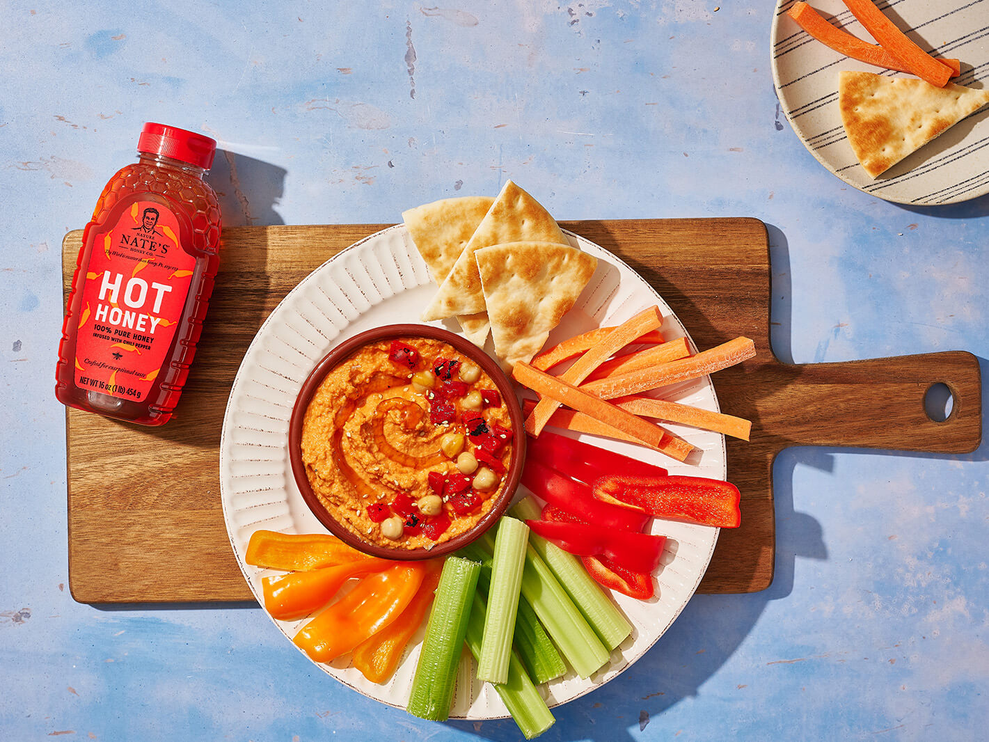 Red pepper hummus dip in a bowl surrounded by veggies and pita on a cutting board with a bottle of Nate's hot honey.