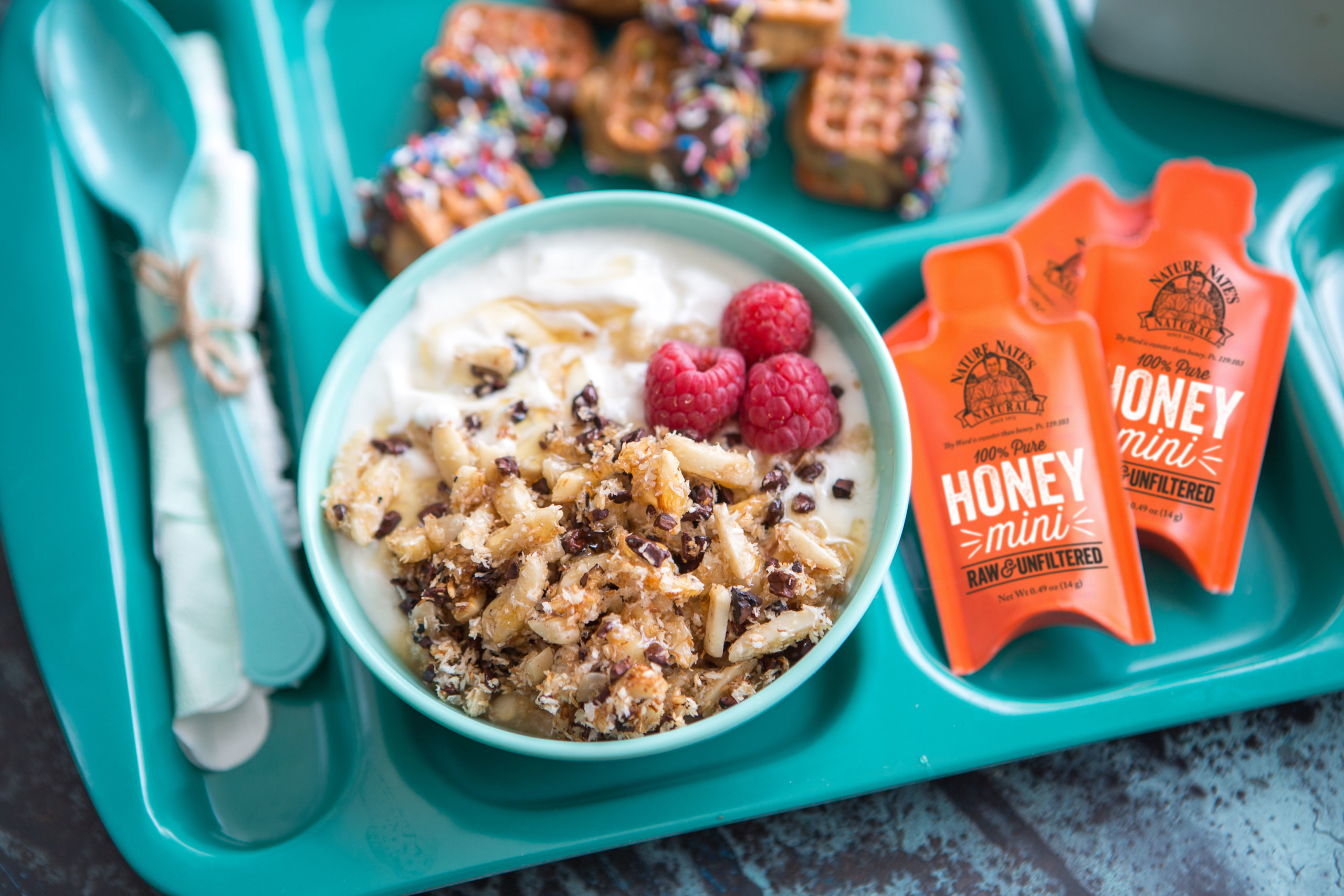 Yogurt bowl topped with almond and coconut crunch and berries, Nate's honey minis on a teal tray with pretzel treats.