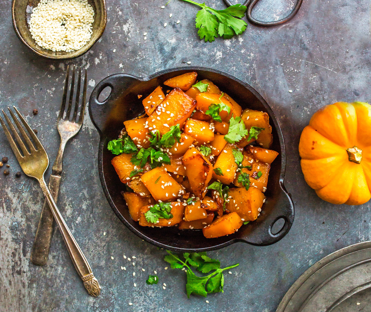 Roasted butternut squash with sesame seeds, herbs, and a small pumpkin on a gray backdrop.