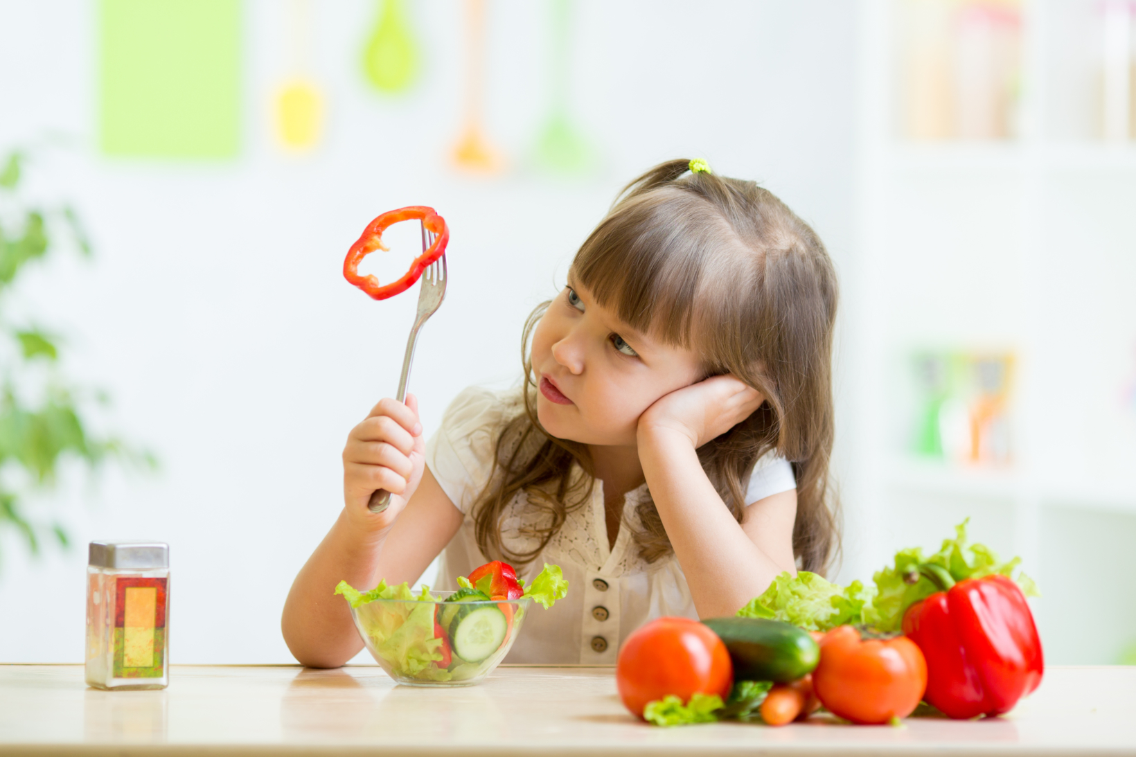 Young girl contemplating a slice of red bell pepper on her fork, surrounded by fresh vegetables.