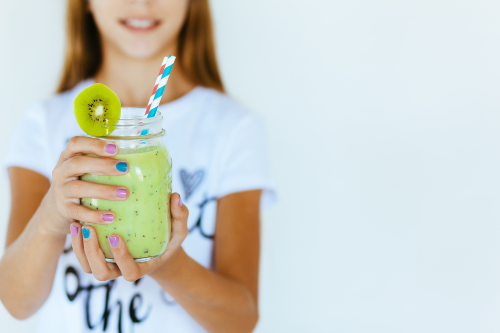 A smiling girl holding a light green smoothie with a colorful straw, in a casual white tee, close-up.