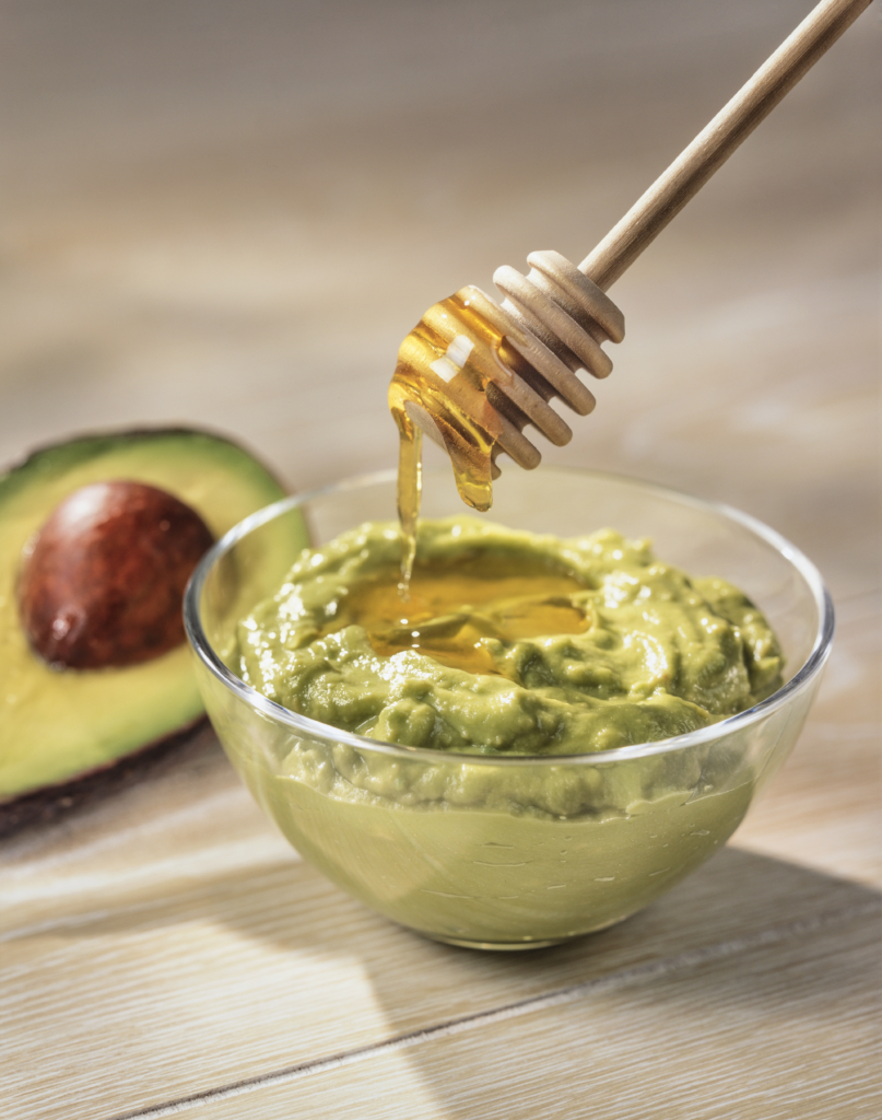 Drizzling honey over avocado dip, with ripe avocado half in the background, on a wooden table.