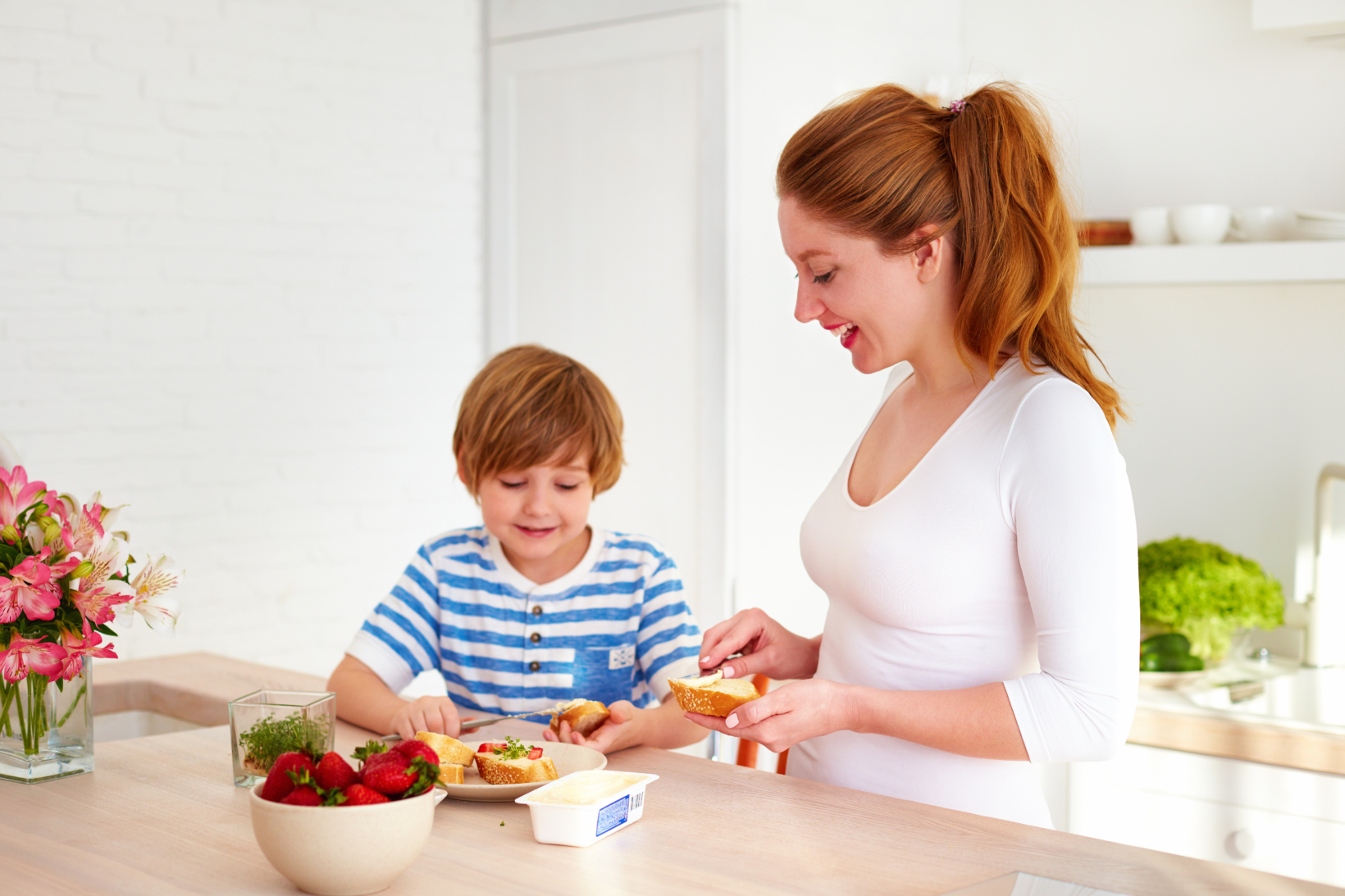 A woman and child prepare food in a sunny kitchen with fresh fruit and flowers.