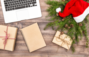 Christmas setting with gifts, Santa hat, and greenery on a wooden surface, alongside a laptop and notepad. Christmas setting with gifts, Santa hat, and greenery on a wooden surface, alongside a laptop and notepad.