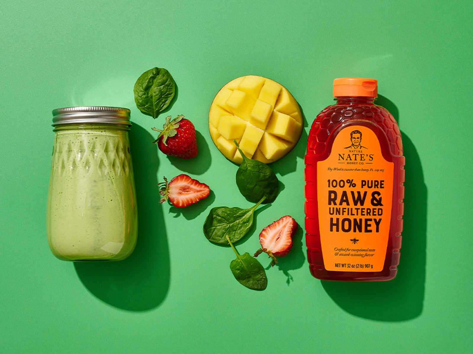 Green smoothie with fruits, spinach, and a bottle of Nate's raw honey on a vibrant green background.