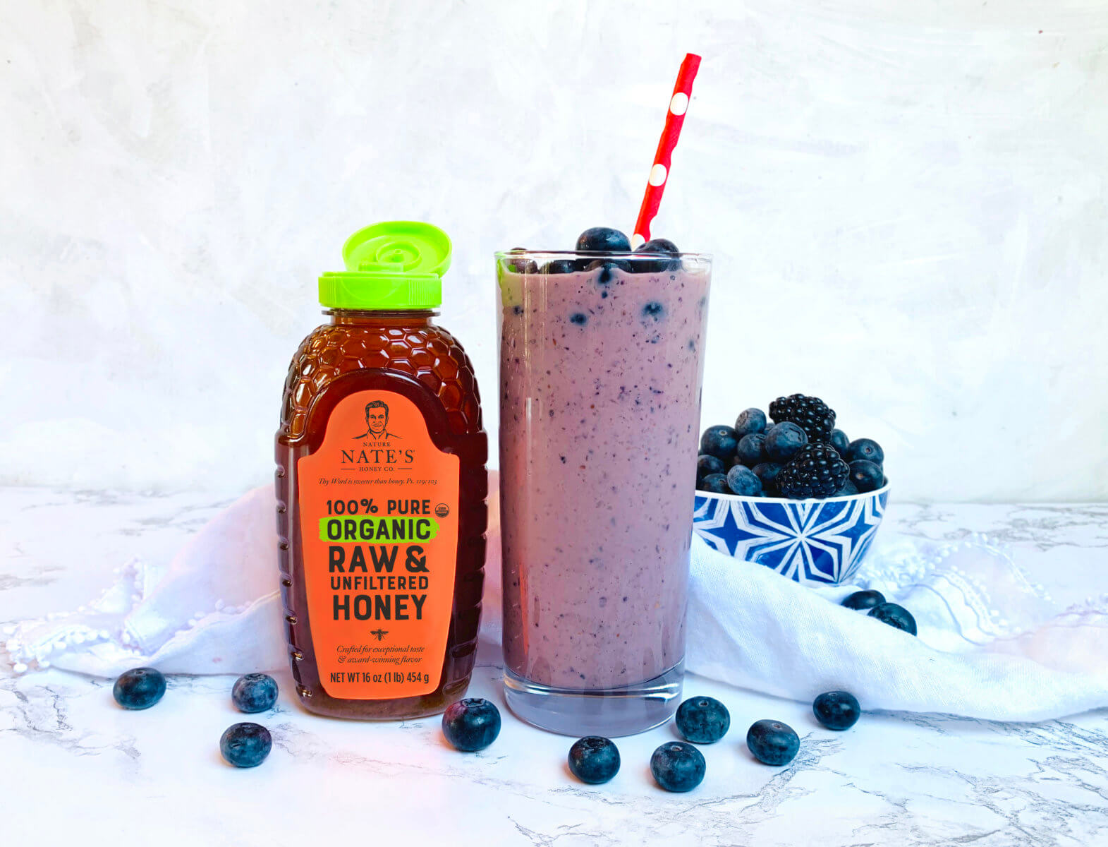 Blueberry smoothie with straw, bottle of Nate's organic honey and bowl of berries on a marbled background.