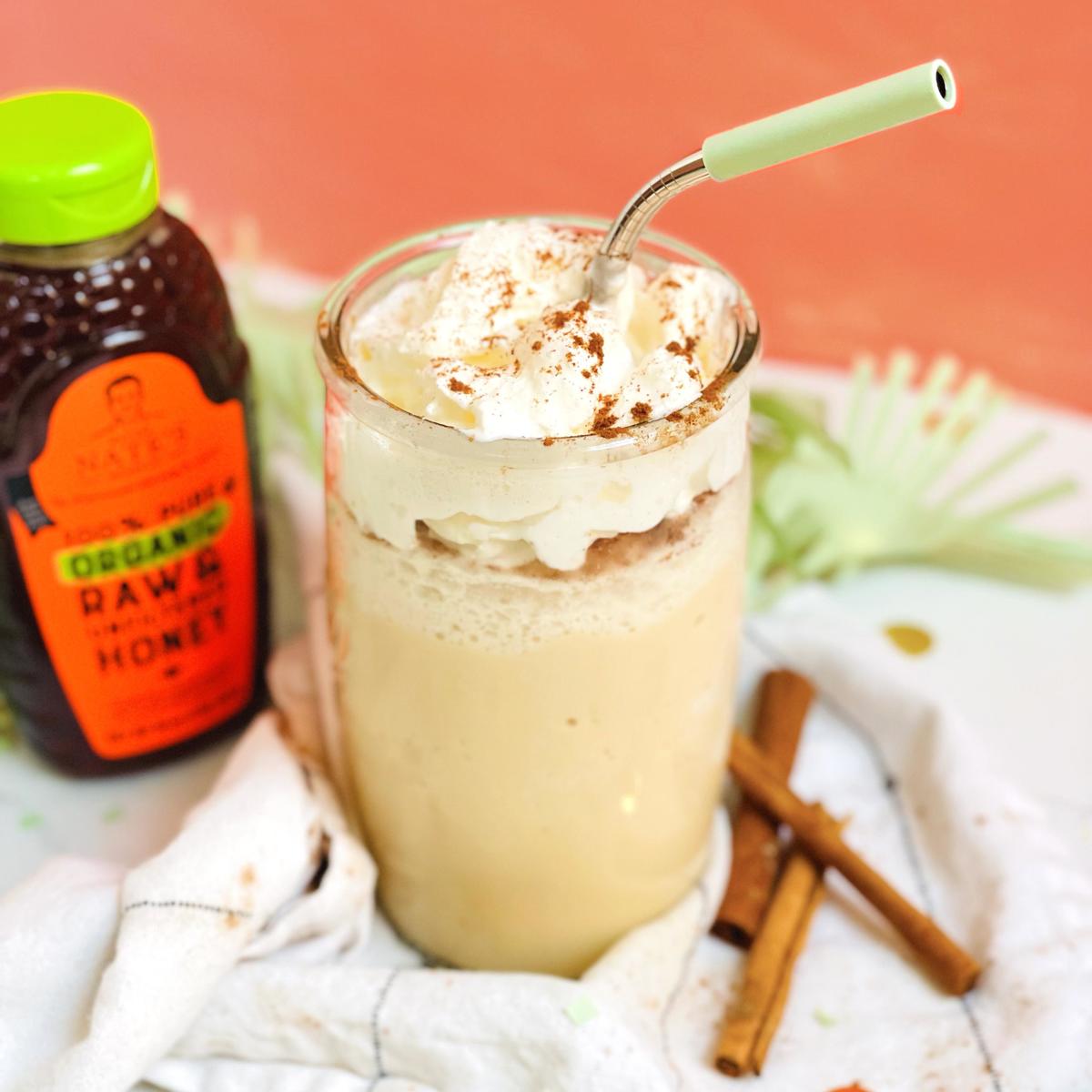 A creamy frappe topped with whipped cream and cinnamon, a bottle of Nate’s organic honey and cinnamon sticks on a cloth.