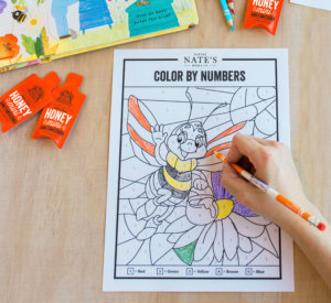 Child's hand coloring a bee on a Color by Numbers activity sheet with crayons and Nate's honey minis nearby.