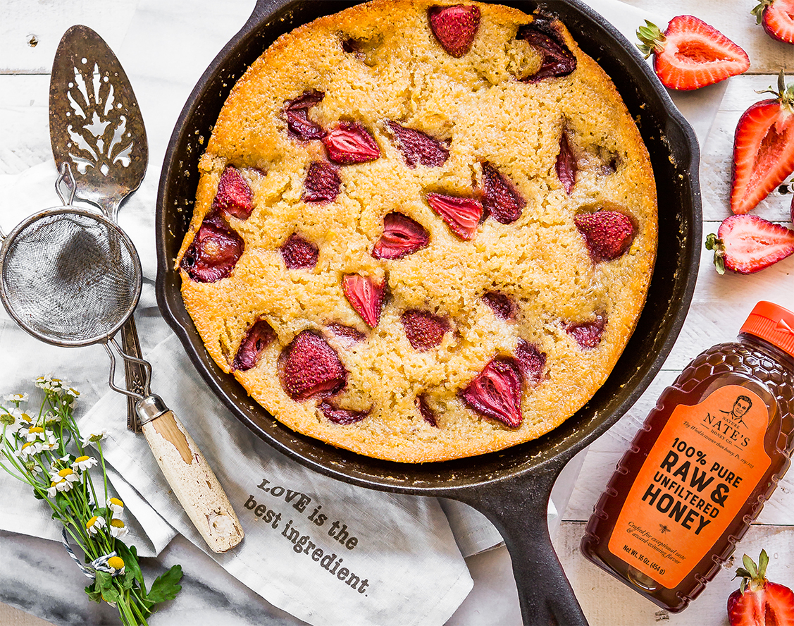 Strawberry corn cake in a skillet with a bottle of Nate's honey and kitchen utensils, with strawberries and herbs.