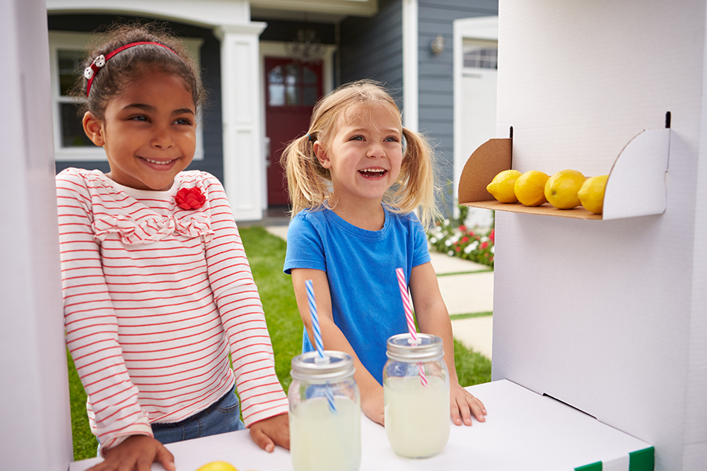 Two smiling children at a lemonade stand with a suburban background.