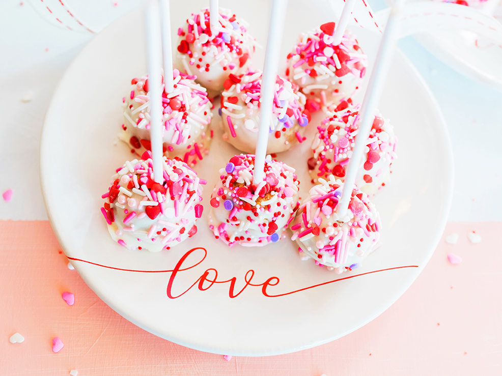 Cake pops with white frosting and pink sprinkles on a plate with love, written on it.