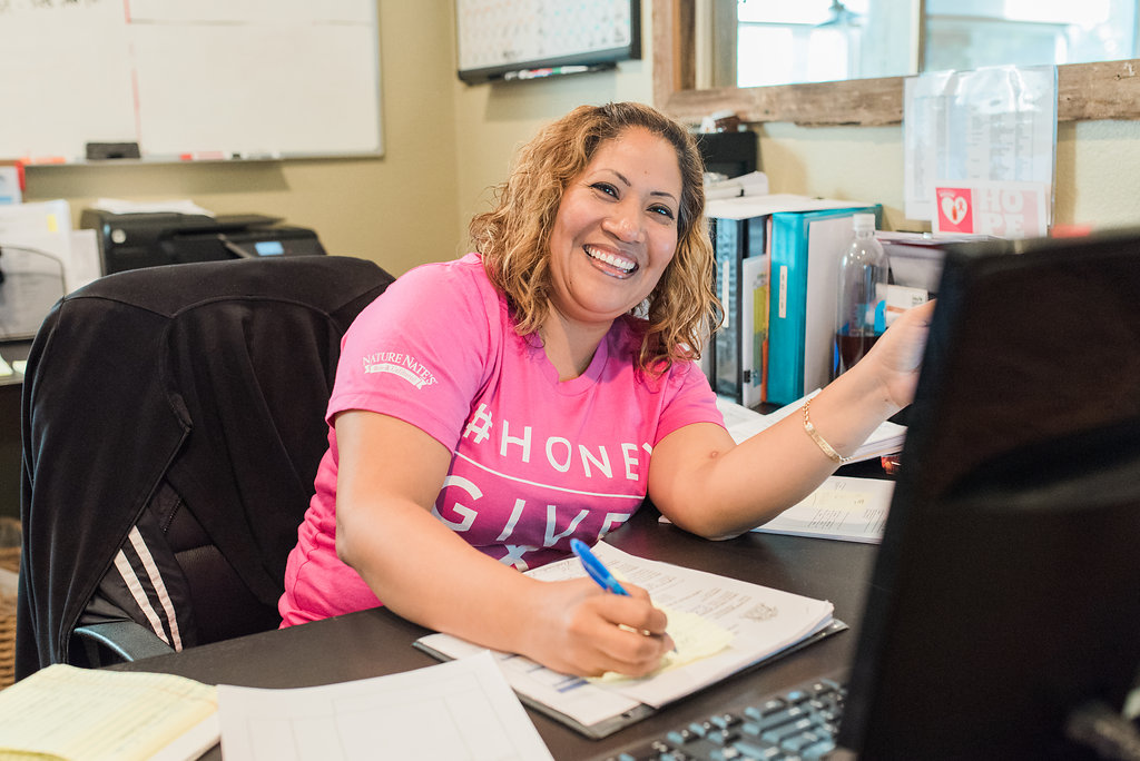 A smiling female employee in a pink shirt with Honey Gives Hope working at a desk with a computer.