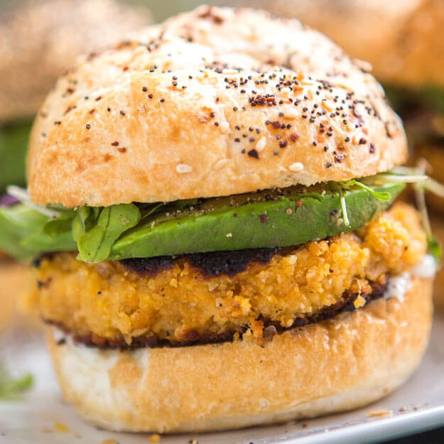 Close-up of a sandwich with a crispy patty, avocado slices, and greens in a seeded bun.