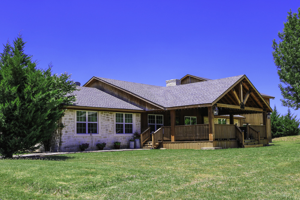A single-story stone house with a prominent wooden gable, front deck, and green lawn, under a clear blue sky.