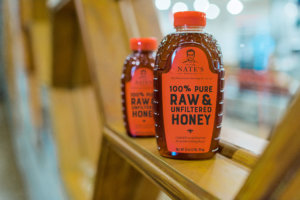 Two bottles of Nature Nate's 100% Pure Raw & Unfiltered Honey on a wooden shelf in a store setting.