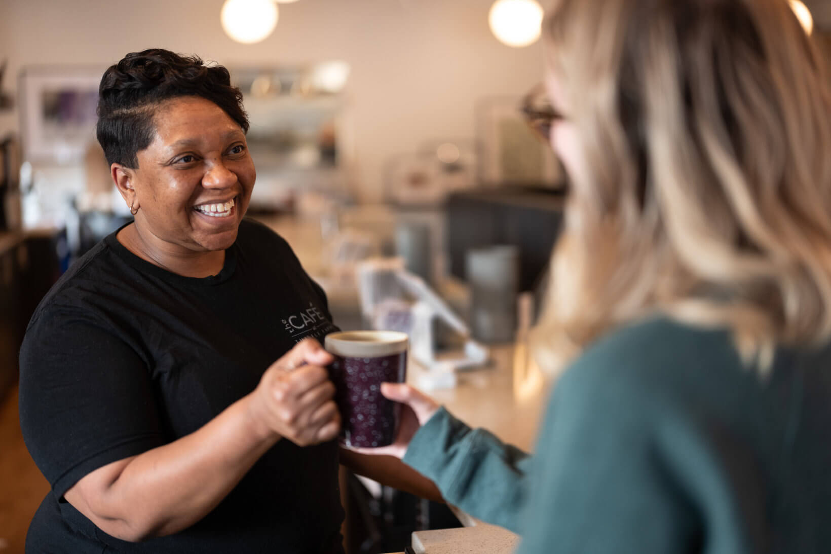 A smiling barista hands a cup of coffee to a customer in a cozy cafe setting.