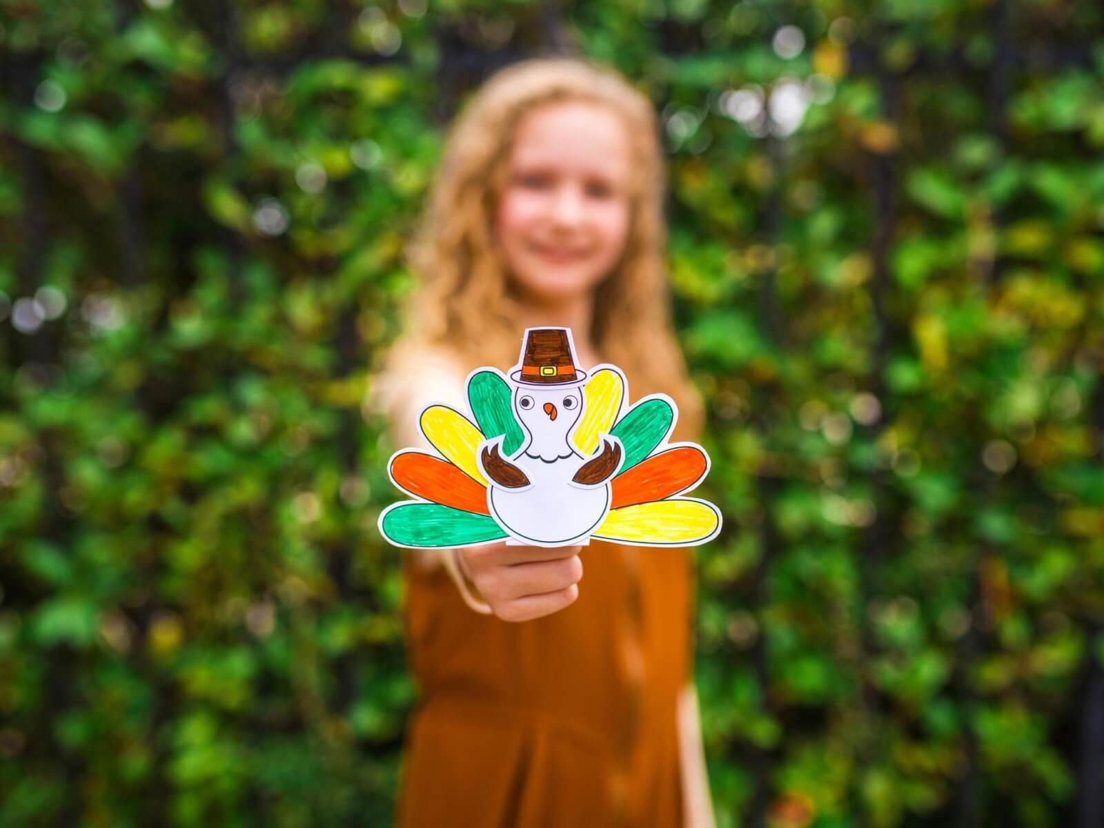 A young girl with curly hair, holding a colorful turkey handcraft, stands in front of a lush green hedge.