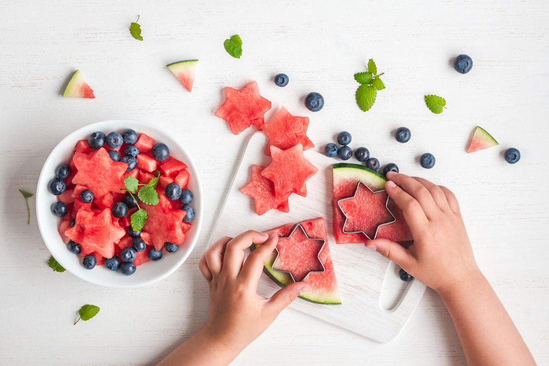 Child making watermelon stars with blueberries and mint on a white table.