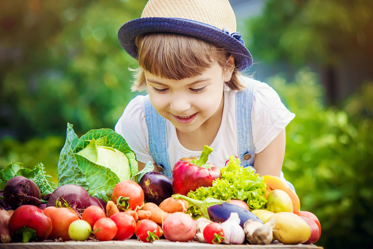 Child in a hat smiling at colorful fresh vegetables and fruits.