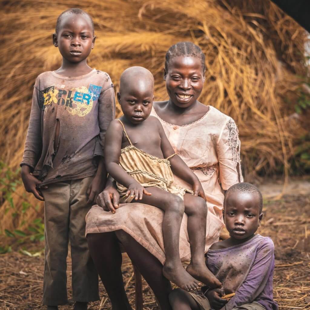 A woman and three children posing with warm smiles, in front of a thatched structure. A woman and three children posing with warm smiles, in front of a thatched structure.
