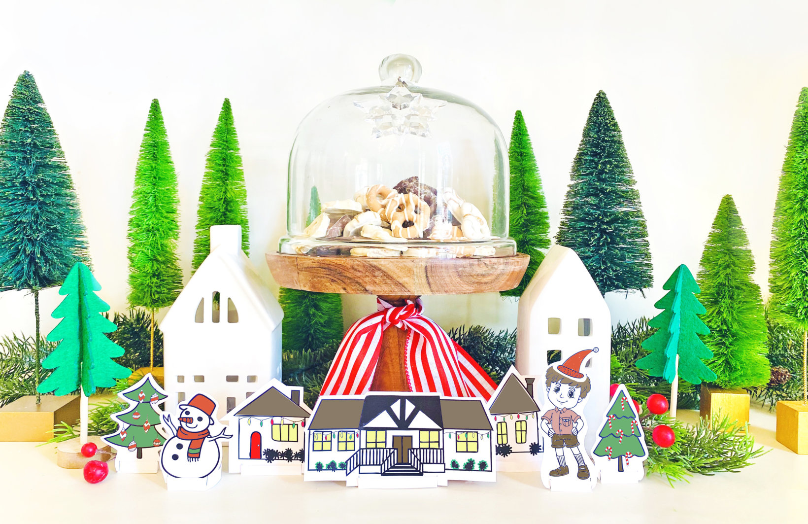 A festive holiday display with paper cutout houses, snowmen, a glass cloche with cookies, green trees, and red ornaments.