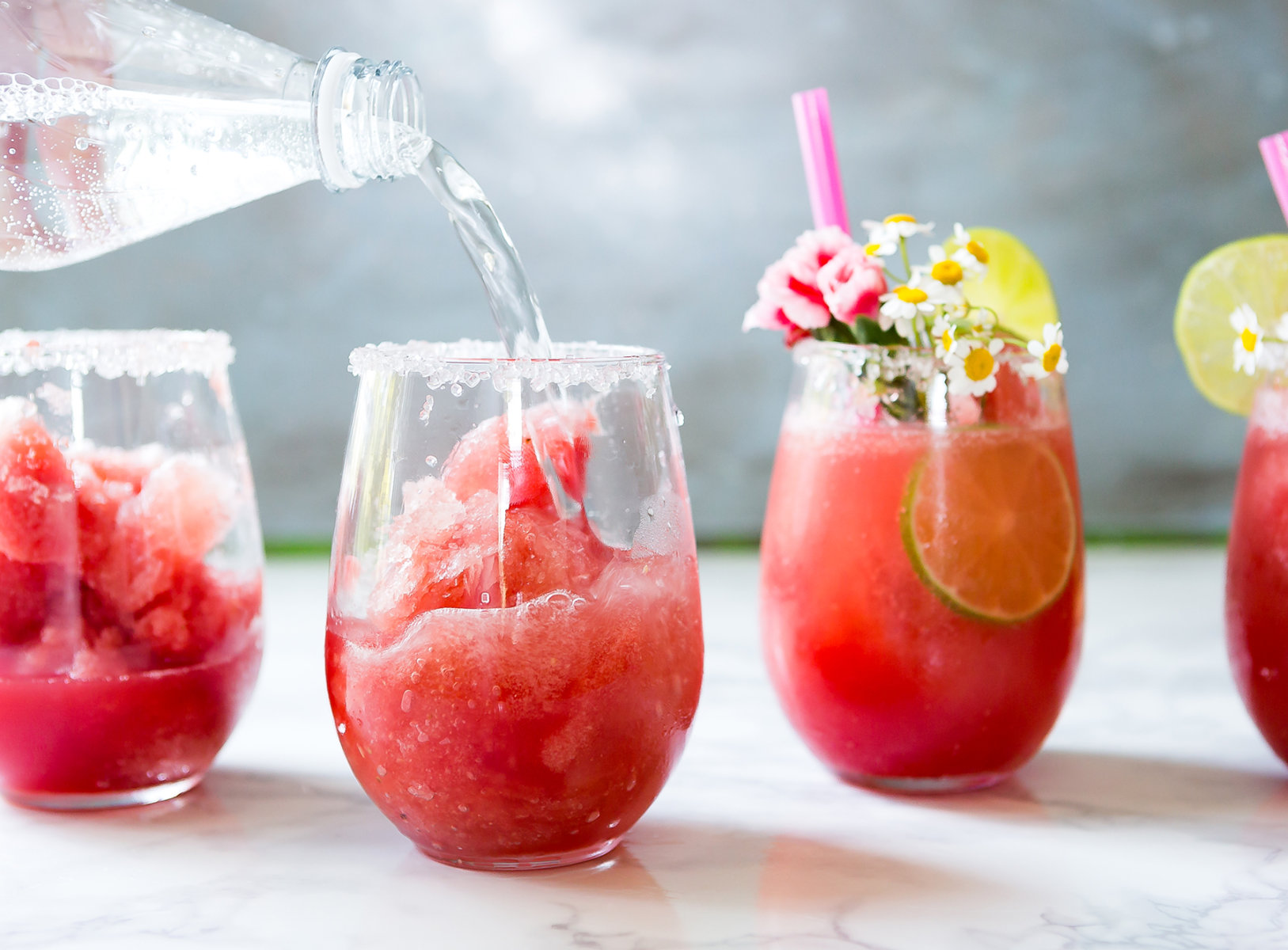 Pouring sparkling water into glasses of watermelon slush garnished with lime and flowers on a marble surface.