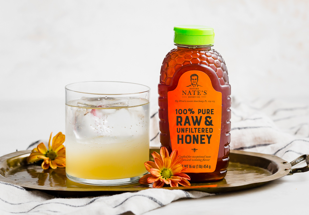 A bottle of Nate's 100% Pure Raw & Unfiltered Honey next to a glass of beverage on a metallic tray with flowers.