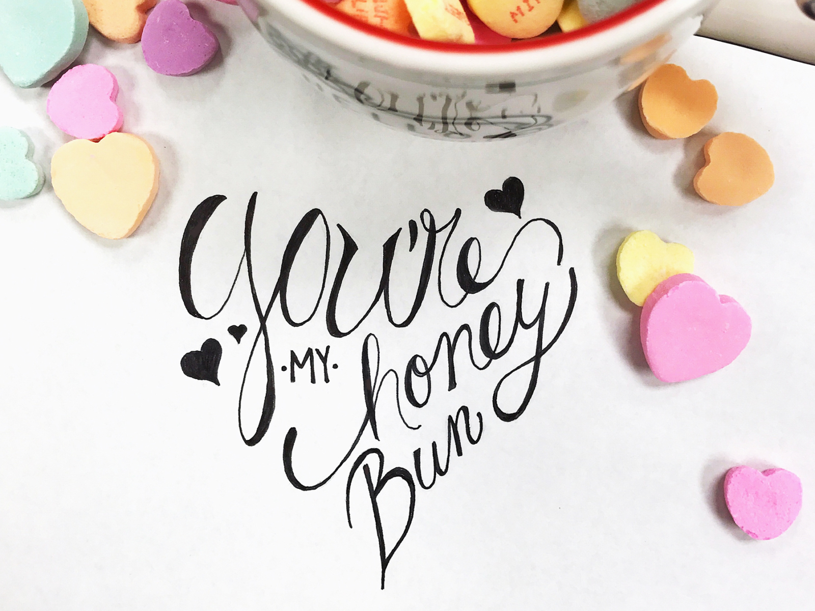 A handwritten note saying You're my honey bun surrounded by colorful heart-shaped candies and a bowl in the background.