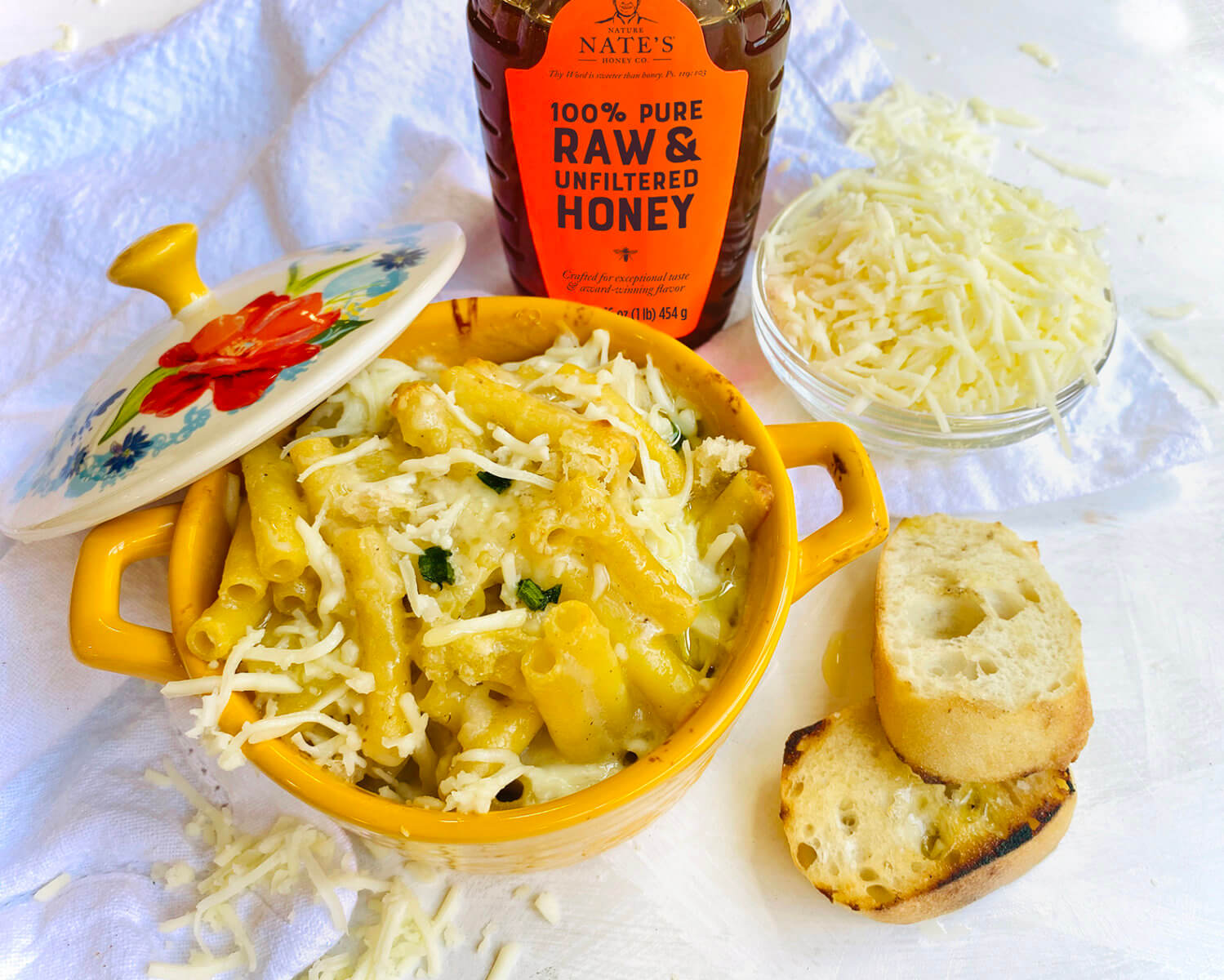 Cheesy pasta in a yellow dish with a bottle of Nate’s honey, shredded cheese and garlic bread on crinkled white cloth.