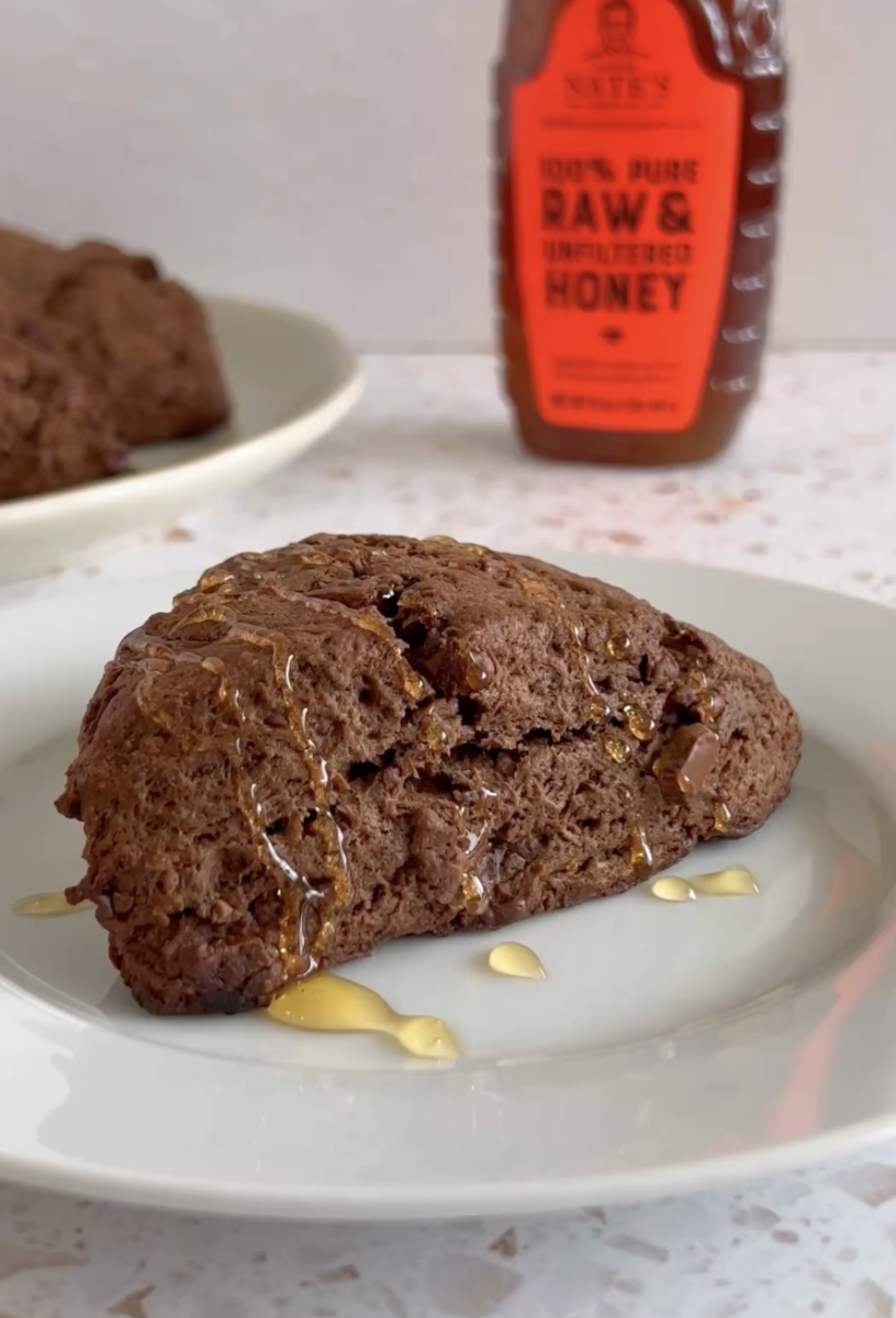 A chocolate scone drizzled with Nate's honey on a plate, with a bottle of Nature Nate's honey in the background.